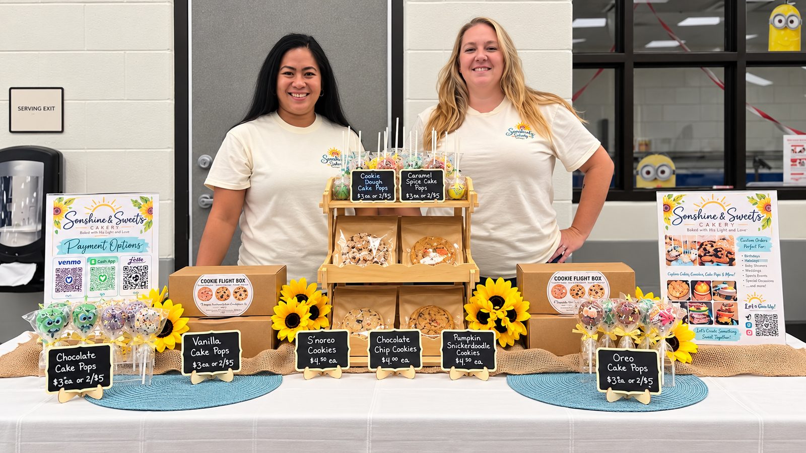 Two smiling bakers behind the Sonshine and Sweets market table, with cookie boxes, cake pops and sunflowers